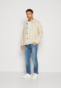 Beige, button-up jacket with four pockets, paired with a white t-shirt, blue jeans, and white sneakers. Smooth fabric texture.