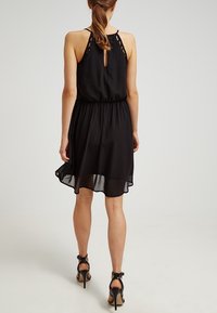 Robe noire dos nu avec une taille cintrée, une jupe fluide et des détails en dentelle au dos. Elle présente une ouverture en forme de goutte d'eau et une texture douce et transparente.