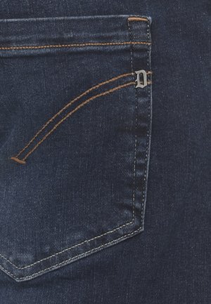 Tasca posteriore di jeans in denim blu scuro con cuciture arancioni e un piccolo ornamento metallico a forma di "D" sulla cucitura superiore destra.