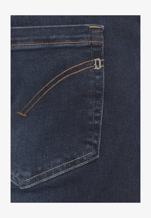 Tasca posteriore di jeans in denim blu scuro con cuciture arancioni e un piccolo ornamento metallico a forma di "D" sulla cucitura superiore destra.