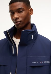 Άνδρας που φοράει μια σκούρη μπλε μπουφάν Tommy Hilfiger με ψηλό γιακά και λευκό πουλόβερ από μέσα, κοιτάζει μπροστά με ουδέτερη έκφραση.