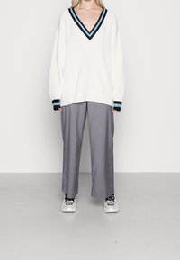 Un pull en tricot blanc avec un col en V et des poignets rayés noirs et bleus, associé à un pantalon gris à rayures et des baskets blanches.