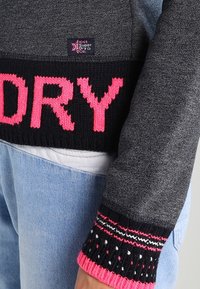 Pull en gris avec un ourlet noir côtelé arborant "DRY" en rose vif. Les manches côtelées présentent un motif tricoté multicolore avec des accents roses.