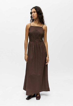 Femme aux longs cheveux bouclés portant une robe longue marron sans manches avec un détail losange brodé, debout devant un fond blanc uni.