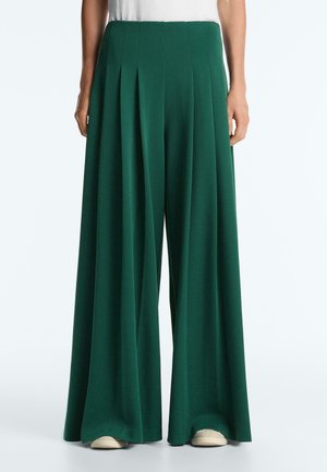 Green wide-leg pleated trousers worn by person standing, paired with white shoes and a white top, against a plain background.