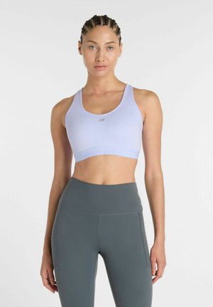 Kvinde med flettet hår iført lys lilla sportstop og grå leggings med høj talje, stående mod en ensfarvet hvid baggrund.