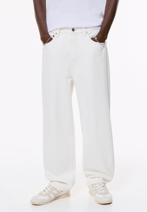Pantaloni ampi in denim bianco con vita alta e tasche laterali, dotati di chiusura a bottone e dettagli di cucitura minima. Abbinati a sneakers chiare.