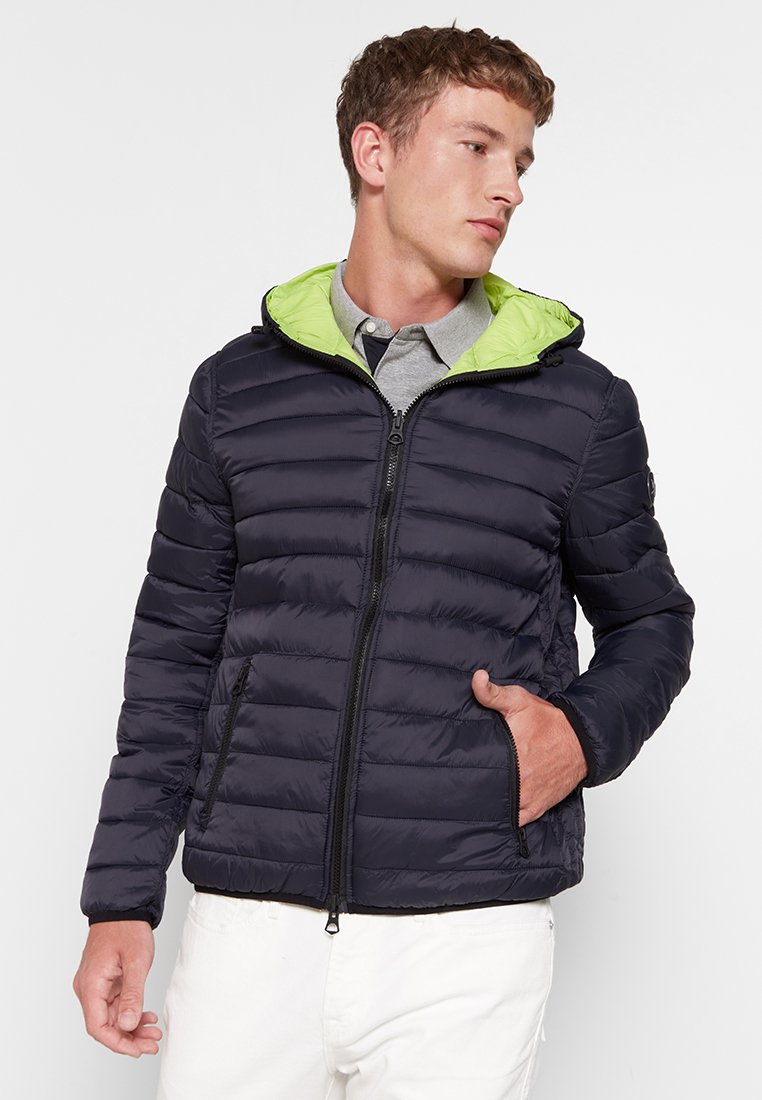 Marineblauwe pufferjack met een lichtgroene capuchon, rits aan de voorkant en zijzakken. Heeft een doorgestikte textuur en een getailleerde pasvorm.