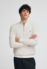 Krämfärgad tröja med zip-up mock-neck, med långa ärmar och en liten svart logotyp. Mjuk, texturerad tyg.