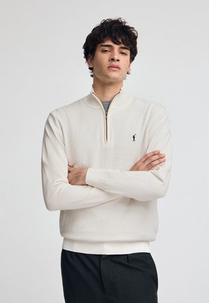 Crèmekleurige pullover met een ritskraag, voorzien van lange mouwen en een klein zwart logo. Zacht, textuur-achtig materiaal.