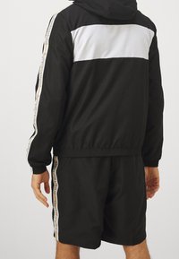 Man som bär en svart och vit huvtröja och svarta shorts med beige detaljer och "LACOSTE" text på ärmarna och sidorna.