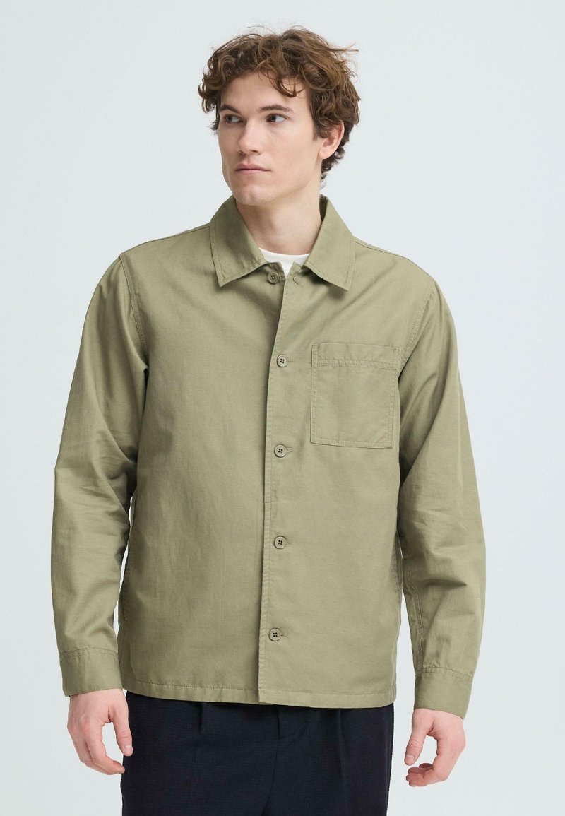 Młody mężczyzna o kręconych włosach, ubrany w zapiętą na guziki oliwkowozieloną koszulę i ciemne spodnie, spoglądający lekko w prawo na jednolitym tle.