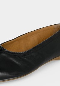 Ballerine en cuir noir avec doublure intérieure beige et un petit nœud près de l’orteil arrondi sur fond blanc.