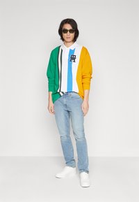 Farbiger Rugby-Shirt mit grünen, gelben, weißen und blauen Streifen, ausgestattet mit einem Kragen und einem gestickten Logo. Getragen mit hellblauen Jeans und weißen Sneakern.