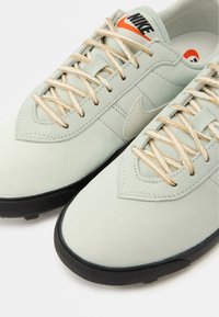 Ανοιχτό γκρι αθλητικά παπούτσια με υφασμάτινο επάνω μέρος, εκρού κορδόνια και μαύρη λαστιχένια σόλα. Διαθέτουν διακριτικό λογότυπο Nike στο πλάι και στη γλώσσα.