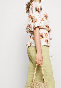 Kremowa bluzka w kwiaty z pomarańczowymi makami, z talią ściśniętą, z krótkimi rękawami, połączona z fakturowanymi oliwkowymi spodniami i okrągłą plecioną torbą.