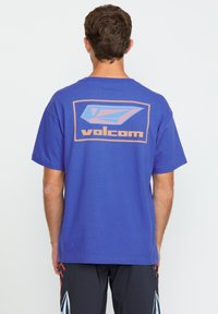 Paarse T-shirt met een grote grafiek in blauw en oranje met de tekst "volcom" op de achterkant. Katoenen materiaal, korte mouwen, relaxte pasvorm.