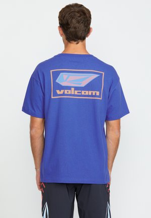 T-shirt violet avec un grand graphique bleu et orange sur le dos, portant le texte "volcom". Matière en coton, manches courtes, coupe décontractée.