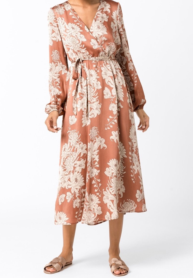 Femme portant une robe midi à manches longues couleur rouille avec un motif floral, une ceinture à la taille et des sandales ouvertes sur fond blanc.