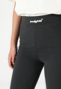 Leggings grigi con una texture morbida e elastica. Caratterizzati da una vita alta con un logo ricamato bianco. Vista ravvicinata della vita e dell'area della coscia.