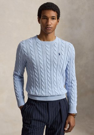 Mand iført lyseblå kabelstrikket sweater og marineblå bukser med nålestriber, stående med hænderne i lommerne mod en neutral baggrund.