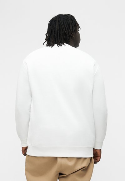 Person mit dunklem geflochtenem Haar, von hinten betrachtet, trägt ein schlichtes weißes Sweatshirt und beige Hosen vor einem hellgrauen Hintergrund.