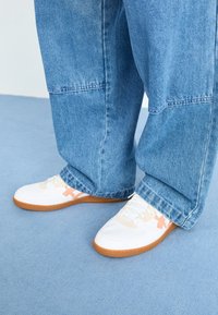 Vita sneakers med gummisulor, med en ljusrosa detalj och en texturerad tygöverdel, bära med löst sittande blå denimbyxor.