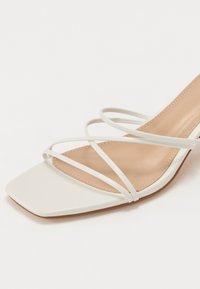 Sandales blanches à talons présentant un bout carré, de fines lanières croisées, et une semelle intérieure beige à texture lisse.