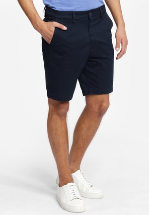 Shorts en coton bleu marine avec devant plat, poches latérales, passants de ceinture et braguette à glissière. Le modèle porte des baskets blanches.