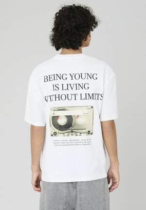 T-shirt blanc présentant un grand graphique d'une cassette audio au dos, avec du texte noir en gras au-dessus et en dessous détaillant la jeunesse et la liberté.