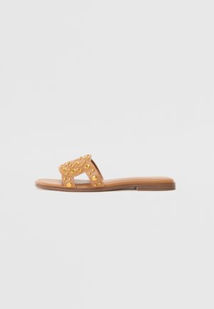 Flache beige Slide-Sandale mit offenem Zehenriemen, verziert mit mehreren runden goldfarbenen Nieten auf einfarbigem Hintergrund.