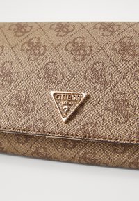Barna textúrájú pénztárca, gravírozott mintával és arany háromszög alakú logoval, amelyen a "GUESS USA" felirat áll. Téglalap alakú, diszkrét fényességgel.