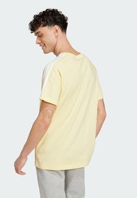 Camiseta de algodón amarilla con rayas blancas en los hombros, corte relajado, escote de cuello redondo y tatuaje visible en el brazo. Combinada con pantalones grises.