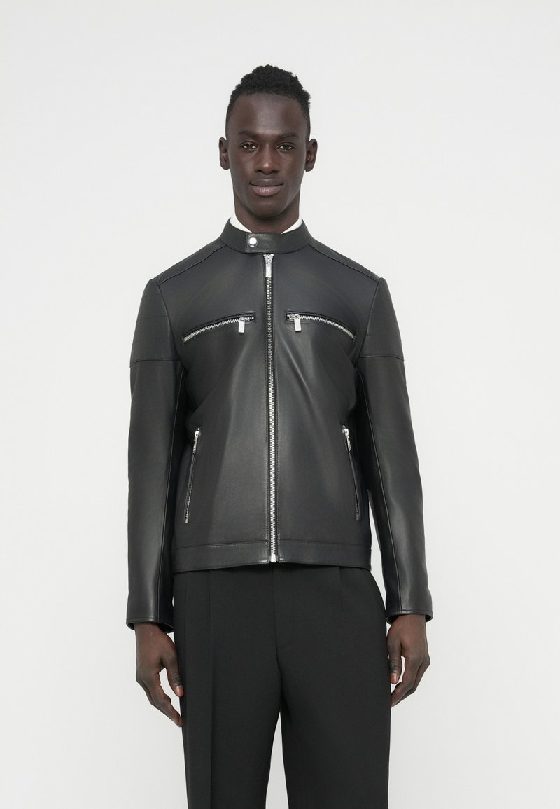 Μαύρο δερμάτινο μπουφάν με ψηλό γιακά, ασημένιες τσέπες με φερμουάρ, ομαλή υφή και εφαρμοστή γραμμή, συνδυασμένο με μαύρο παντελόνι.