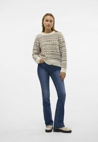 Gestrickter cremefarbener Pullover mit braunen geometrischen Mustern, rundem Ausschnitt und überschnittenen Schultern, kombiniert mit blauen Schlagjeans und cremefarbenen Schuhen.