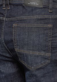 Mörkblå denimjeans med en bakficka, synlig sömnad i ljusbrunt och en rektangulär läderetikett vid midjan.