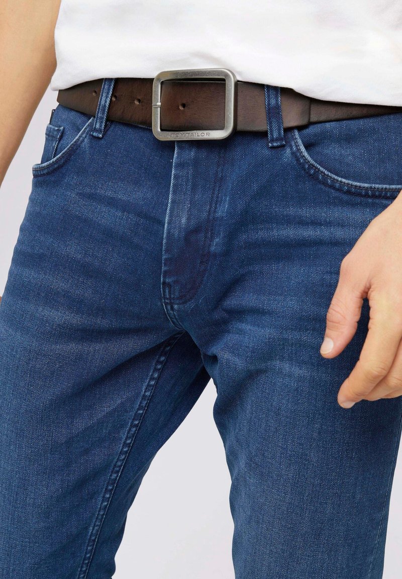 Jeans en denim bleu foncé au design ajusté, complétés par une ceinture en cuir marron avec une boucle métallique carrée.