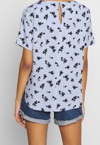 Světlomodrá krátký rukáv halenka s černým květinovým vzorem. Na zádech je výřez ve tvaru klíčové dírky se zapínáním na knoflík. Kombinováno s džínovými šortkami.