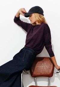 Pull à carreaux violets, jean noir taille haute, casquette en velours côtelé gris. Assis sur une chaise en métal avec un siège en brun texturé.