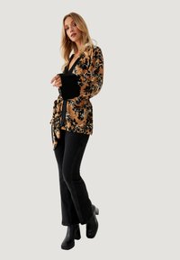 Top de terciopelo marrón con estampado floral, detalles en negro y mangas amplias, combinado con jeans de campana negros y botas de tacón grueso.
