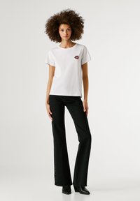 Γυναίκα με σγουρά μαλλιά, φορώντας λευκό μπλουζάκι με μικρό σχέδιο χείλους, μαύρα φαρδιά τζιν με κουμπιά στο πλάι και μαύρες γόβες, στέκεται.