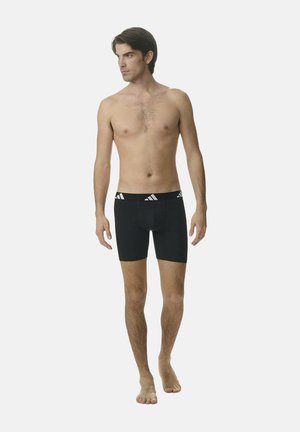 Modelo masculino de pie, descalzo, vistiendo calzoncillos boxers negros con una cinturilla negra que presenta un logo blanco, frente a un fondo claro y liso.