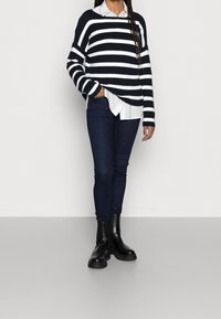 Námořnický a bílý pruhovaný pletený svetr vrstvený přes bílou košili, v kombinaci s tmavými skinny džíny a lesklými černými kotníkovými botami.