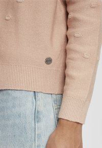 Maglione lavorato a maglia rosa chiaro con dettagli a pallini testurizzati, polsini e orlo a costine, e un accento con logo metallico vicino al fondo.