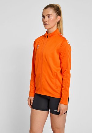 Vrouw met blond paardenstaart, gekleed in een oranje sportjack met rits en zwarte sportshorts, staand tegen een effen lichte achtergrond.