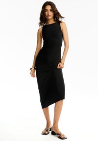 Schwarzes Midi-Kleid aus elastischem Stoff, mit einem taillierten Oberteil, asymmetrischem Saum und gerafften Details. Getragen mit schwarzen Sandalen.