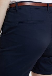 Närbild av en person som bär marinblå shorts med en bakt pocket och ett brunt texturerat läderbälte.