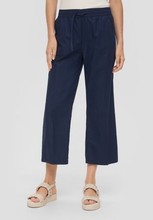 Femme portant un pantalon large court bleu marine avec une taille élastique et un cordon de serrage, assorti à des sandales à brides tressées beige.
