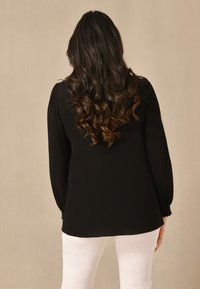 Svart långärmad blus med texturerad tyg och rynkade detaljer vid ärmarna, kombinerad med vita byxor, sedd bakifrån.