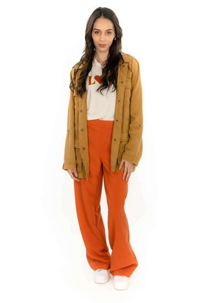 Veste marron à boutons, t-shirt blanc avec un cœur rouge, pantalon orange à jambes larges et chaussures blanches. Textures lisses et couleurs vives.